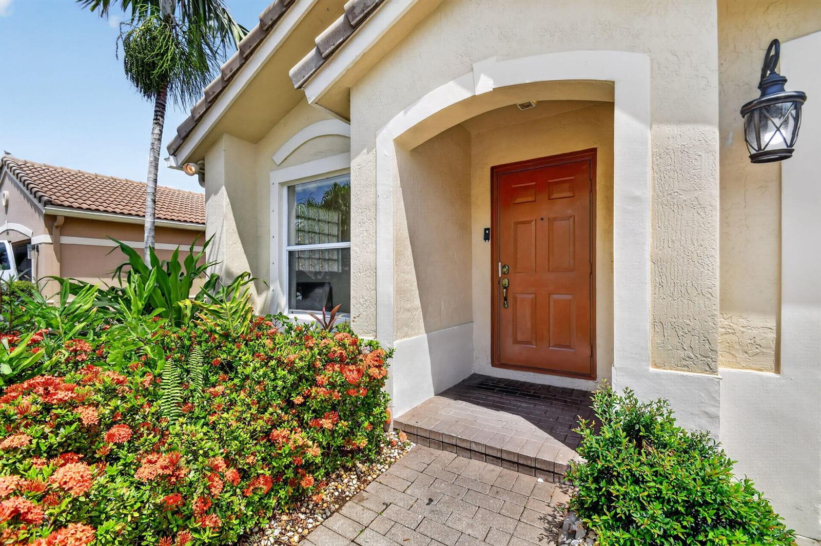 A front entrance with a brown door, brick walkway, and orange flowering shrubs — 1107 Big Torch Street, Riviera Beach FL 33407, RIVIERA BEACH, FL