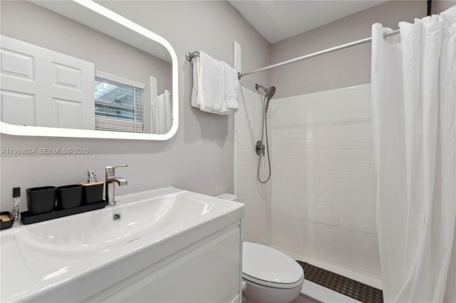 A bathroom features a white vanity, a lighted mirror, and a tiled shower area — 1111 SW 5th St, Boca Raton FL 33486, BOCA RATON, FL