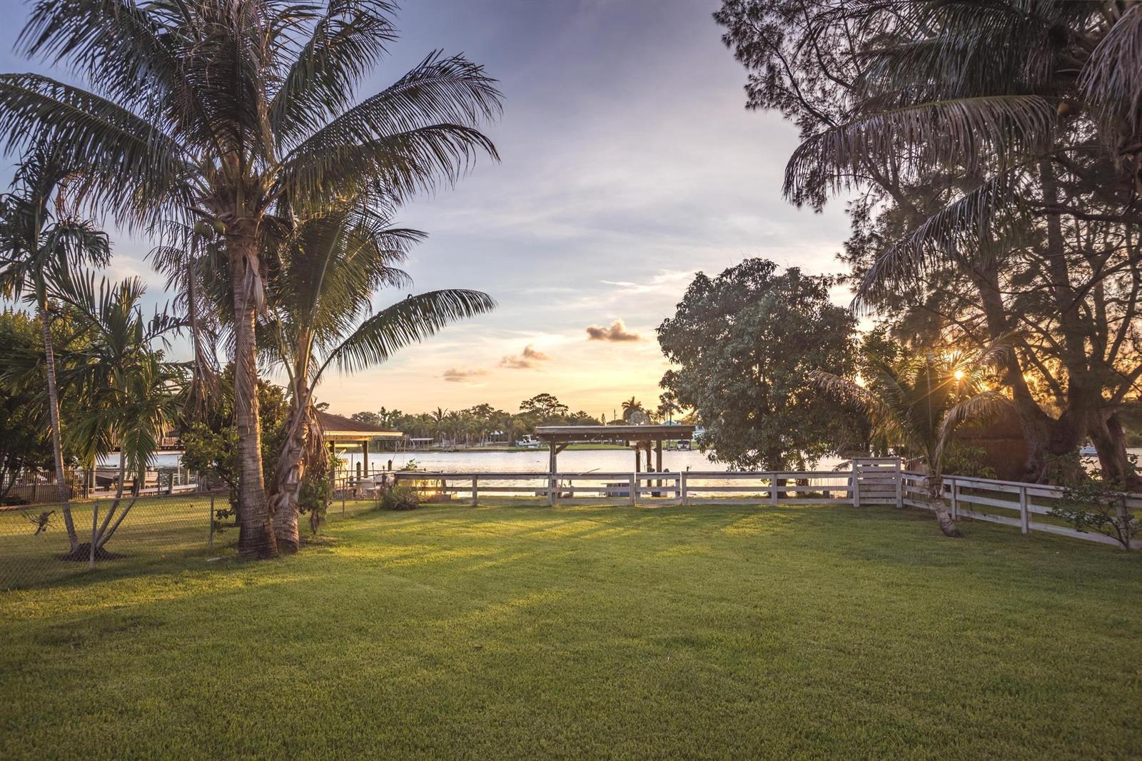 A grassy backyard features a white fence, palm trees, and a view of the water — 6563 Paul Mar Drive, Lake Worth FL 33462, LAKE WORTH, FL