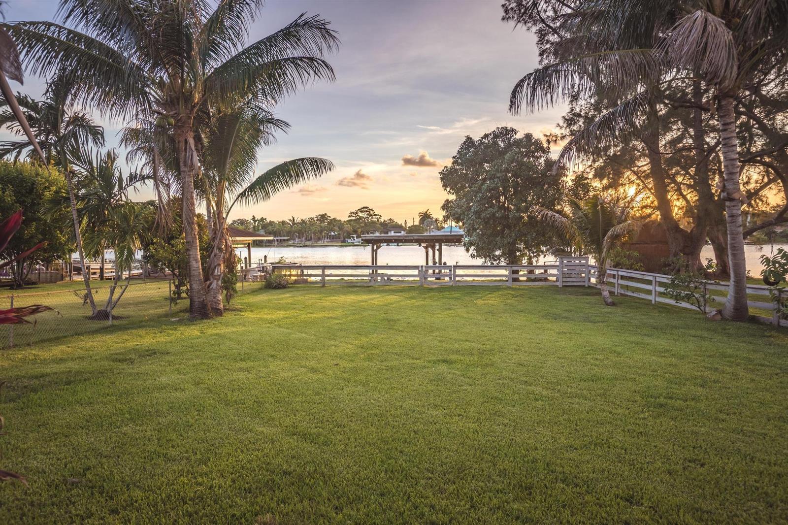 A grassy backyard features palm trees, a white fence, and a view of the water — 6563 Paul Mar Drive, Lake Worth FL 33462, LAKE WORTH, FL