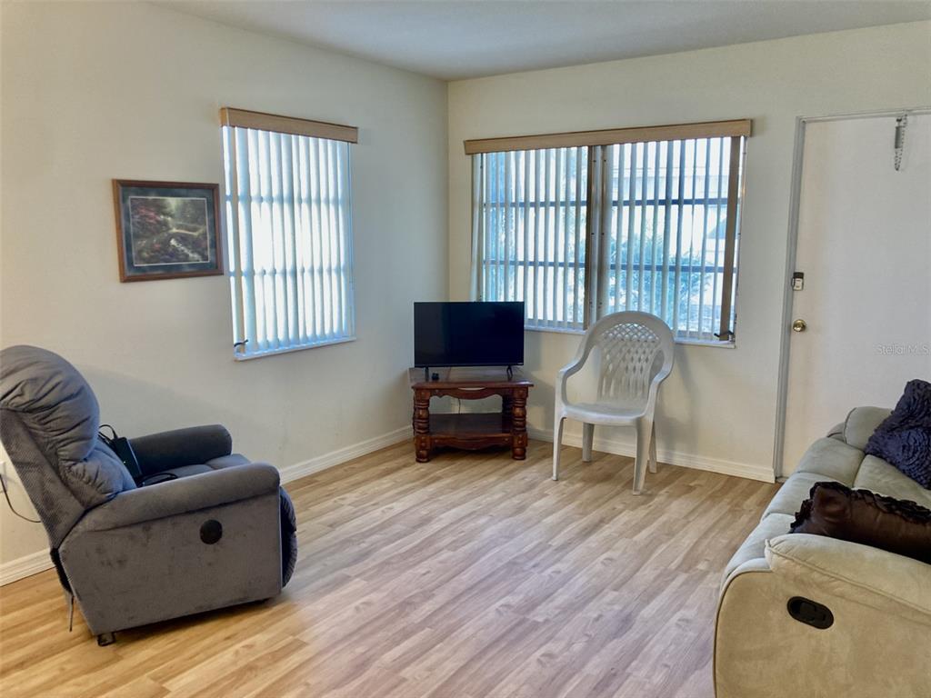 A living room features light wood-look flooring, two windows with vertical blinds, and assorted furniture — 6110 CORAL WAY, BRADENTON, FL