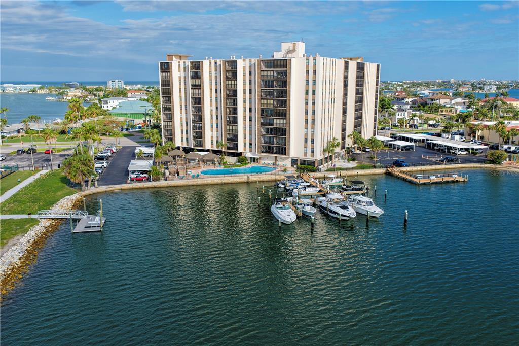 An aerial view shows a multi-story apartment building with a pool and boat docks — 10355 PARADISE BOULEVARD UNIT 907, TREASURE ISLAND, FL