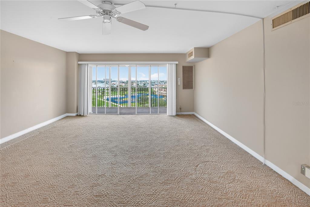 An empty living room features beige carpeting, a ceiling fan, and sliding glass balcony doors — 10355 PARADISE BOULEVARD UNIT 907, TREASURE ISLAND, FL