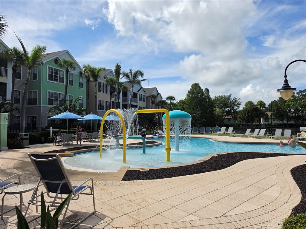 A swimming pool with a splash pad feature sits in front of multi-story apartment buildings — 3016 BONFIRE BEACH DRIVE UNIT 103, KISSIMMEE, FL