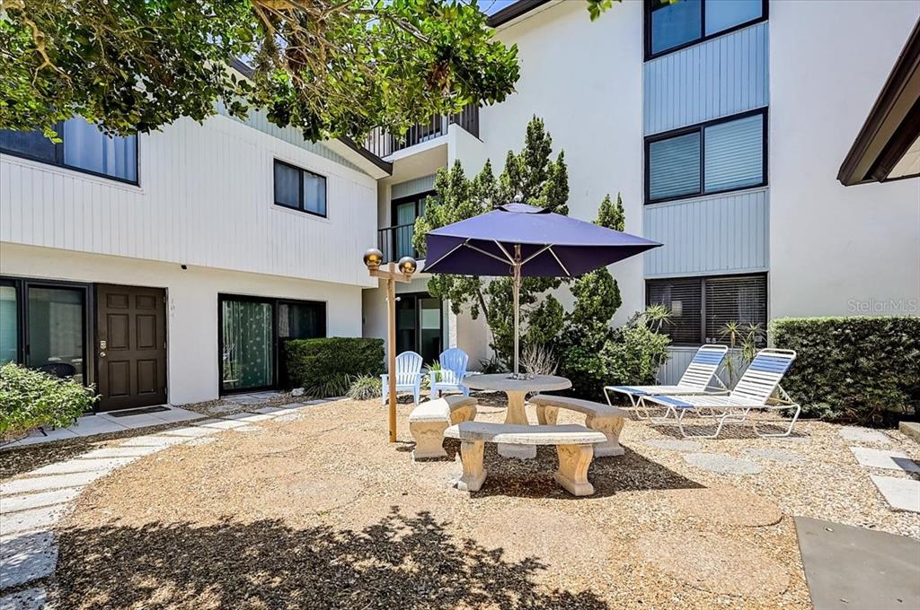 A gravel courtyard features a stone table, umbrella, chairs, and lounge chairs between building wings — 722 S BLVD OF THE PRESIDENTS UNIT 105, SARASOTA, FL