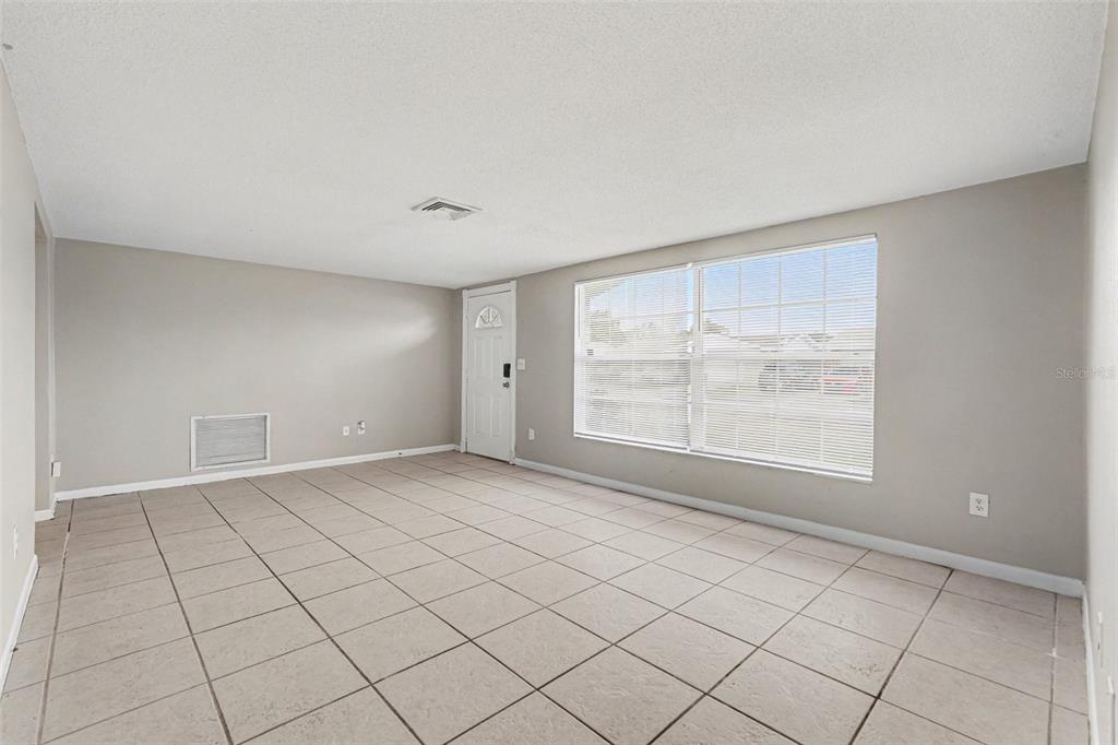 An empty living room features light-colored tile flooring, gray walls, and a large window — 7224 INGLESIDE DRIVE, PORT RICHEY, FL
