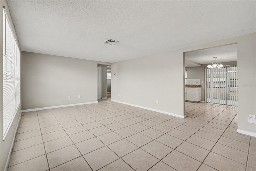 An empty living room with beige tile flooring, light gray walls, and a kitchen beyond — 7224 INGLESIDE DRIVE, PORT RICHEY, FL