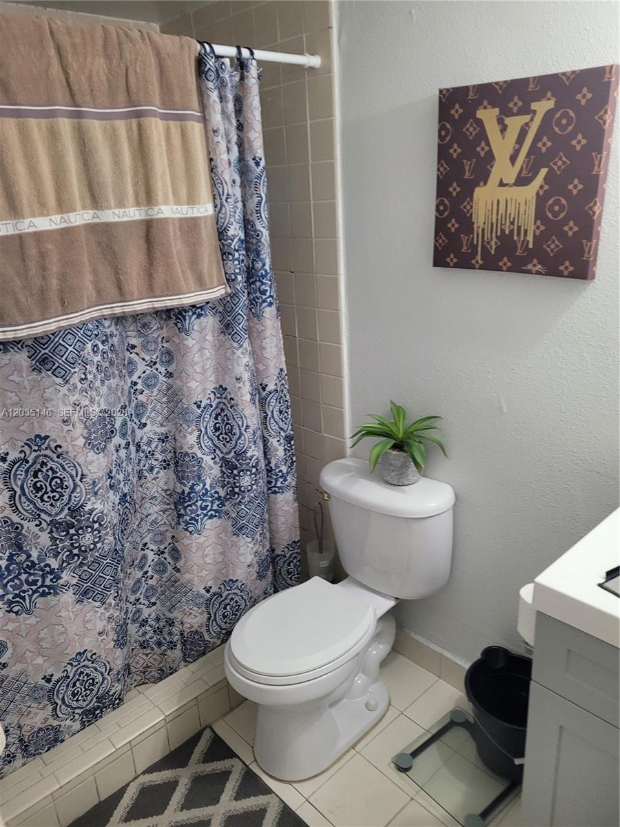 A bathroom features a patterned shower curtain, a toilet with a plant, and wall art — 18101 NW 68th Ave # E105, Hialeah FL 33015, HIALEAH, FL