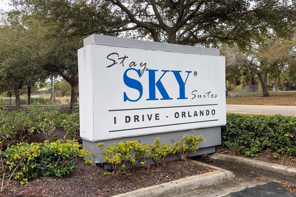 A white rectangular sign for Stay Sky Suites stands outdoors surrounded by trees and bushes — 7601 CANADA AVENUE UNIT 408, ORLANDO, FL