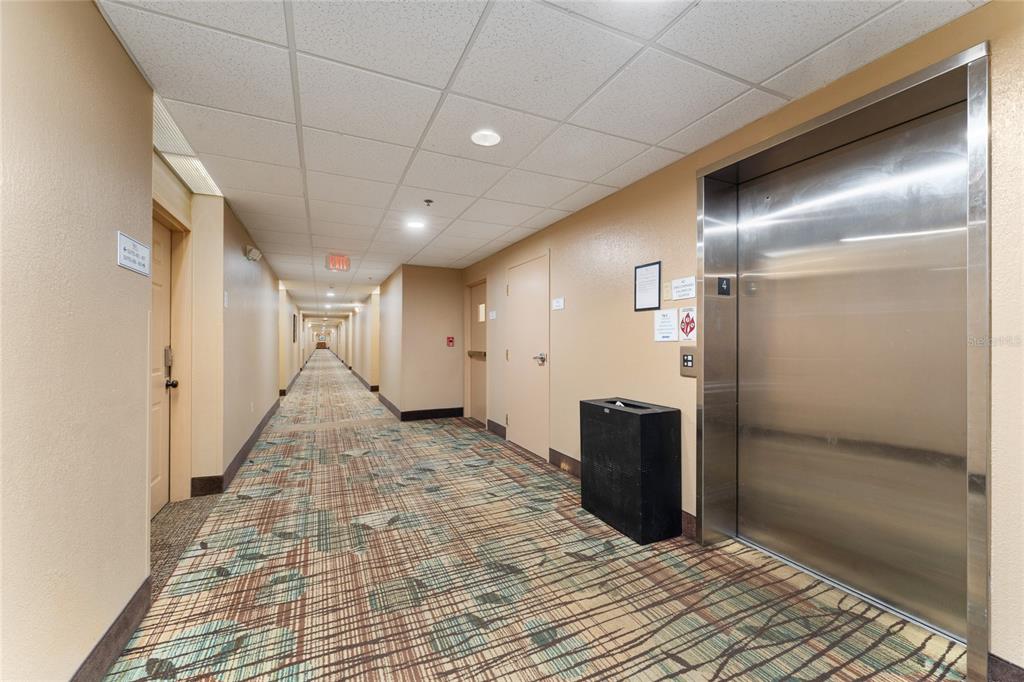 A long hotel hallway features patterned carpet, beige walls, and a stainless steel elevator — 7601 CANADA AVENUE UNIT 408, ORLANDO, FL