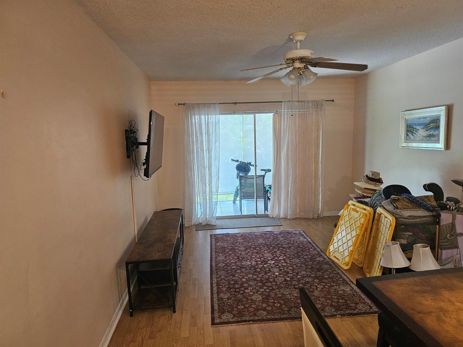A living room features wood-look flooring, a ceiling fan, a wall-mounted television, and sliding glass doors — 2475 W Golf Boulevard # 243, Pompano Beach FL 33064, POMPANO BEACH, FL