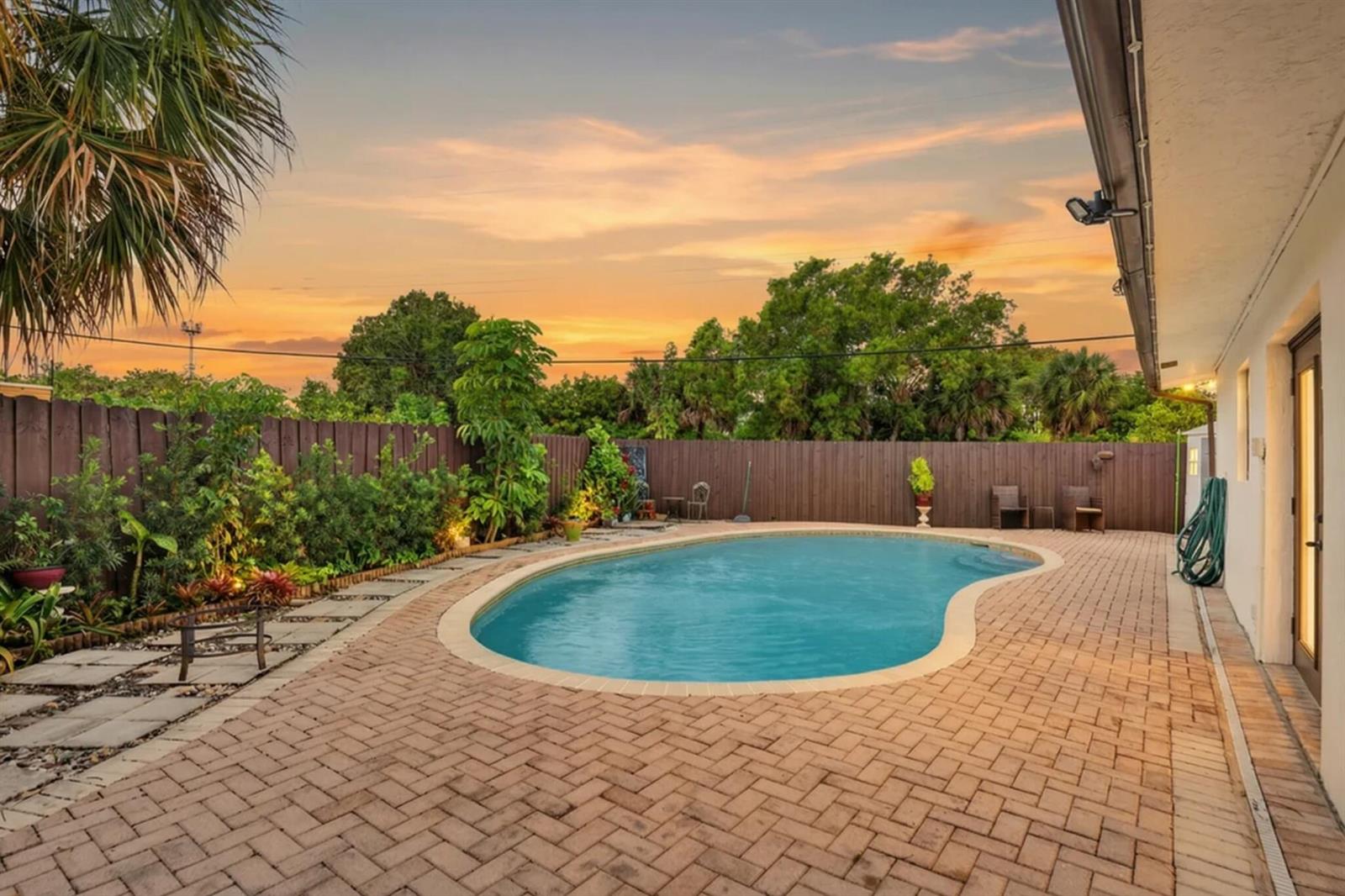 A backyard features a kidney-shaped swimming pool surrounded by brick pavers and a wooden fence — 5051 NE 6th Avenue, Deerfield Beach FL 33064, DEERFIELD BEACH, FL