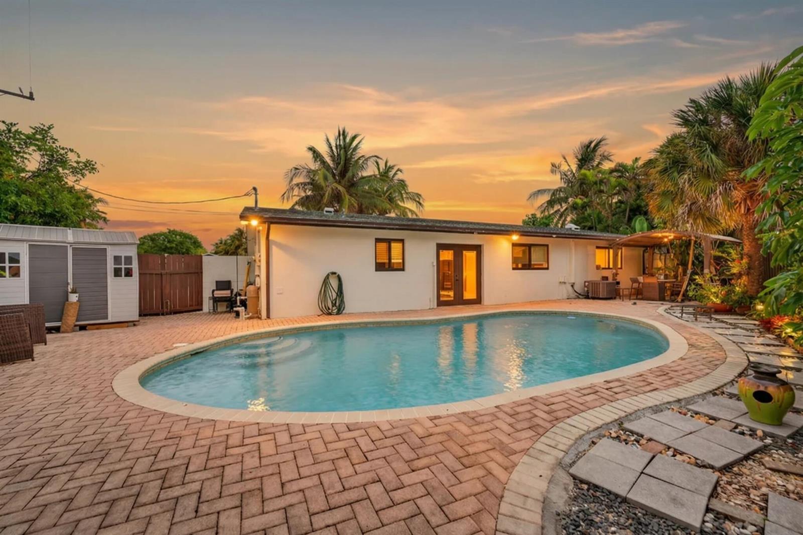 A backyard features a kidney-shaped swimming pool, brick paver patio, and a white house exterior — 5051 NE 6th Avenue, Deerfield Beach FL 33064, DEERFIELD BEACH, FL