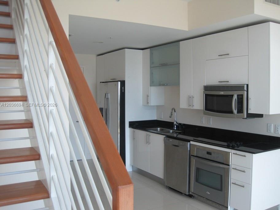 A kitchen with white cabinets, black countertops, and stainless steel appliances sits beside a staircase — 60 SW 13th St # 4610, Miami FL 33130, MIAMI, FL