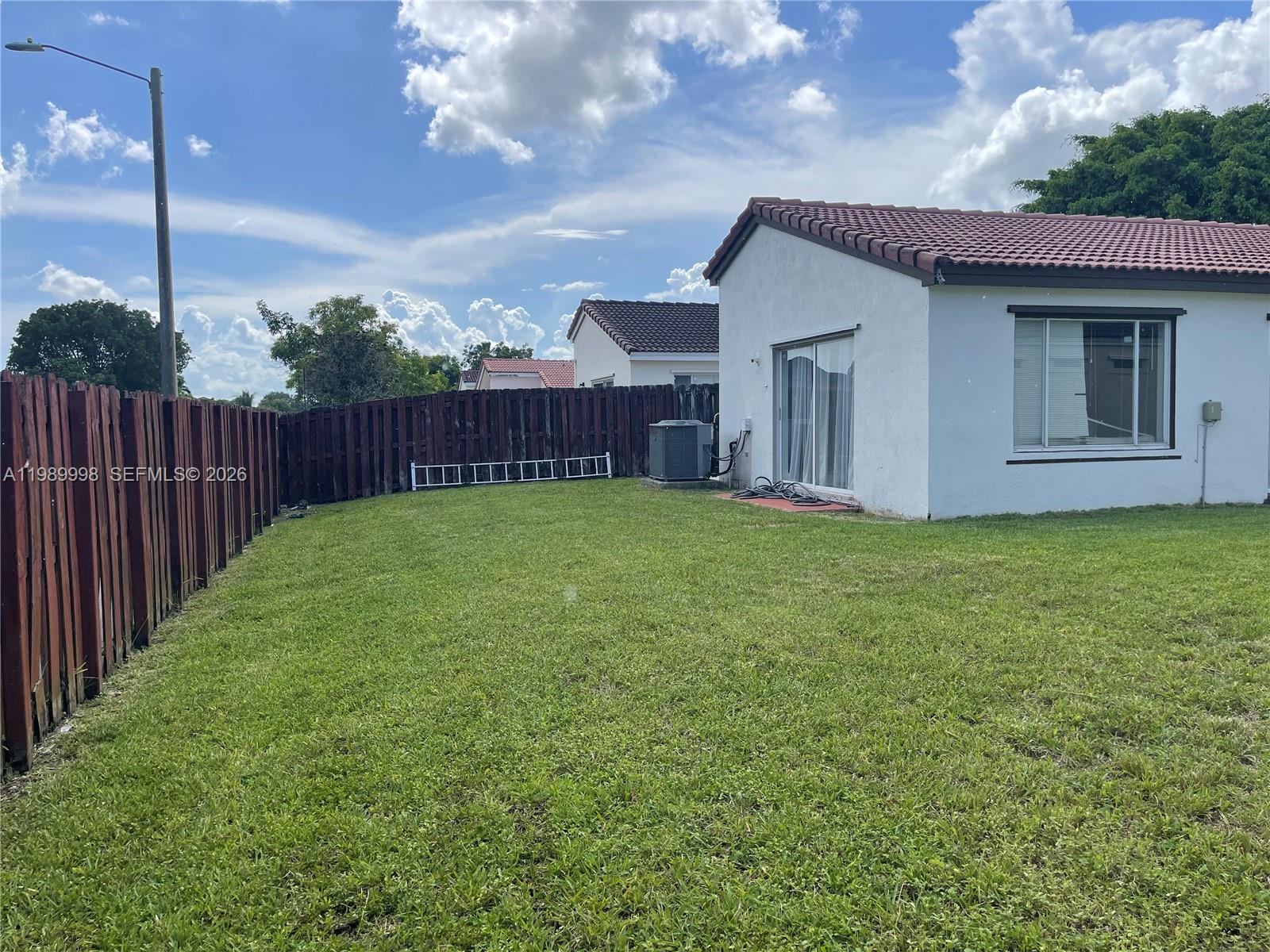 A grassy backyard features a white house with a red tile roof and wooden fencing — 14252 SW 175th Ter, Miami FL 33177, MIAMI, FL