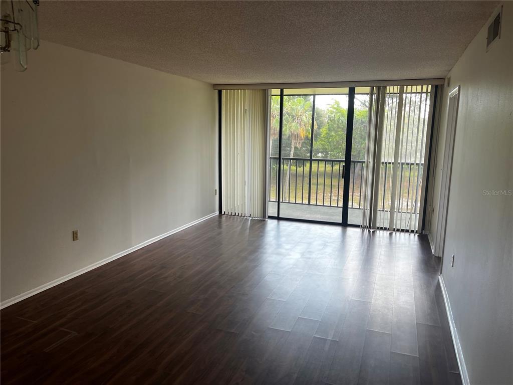 An empty living room features dark wood-look flooring, neutral walls, and a sliding glass balcony door — 800 COVE CAY DRIVE UNIT 3F, CLEARWATER, FL