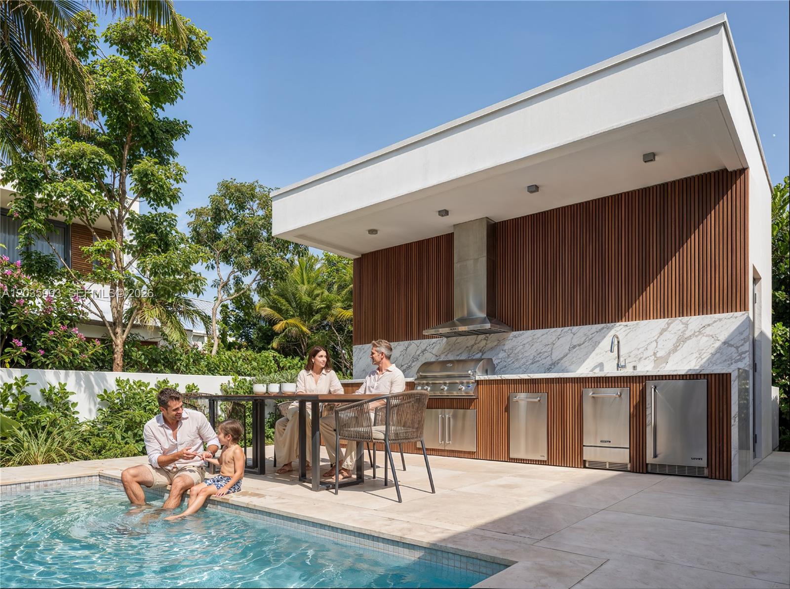 A family sits by a pool next to an outdoor kitchen with marble countertops and wood-slat cabinetry — 9445 SW 73rd Ave, Pinecrest FL 33156, PINECREST, FL
