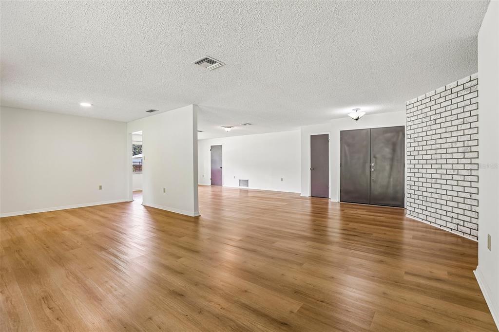 A spacious, open-concept living area features light wood-look flooring, white walls, and a white brick accent wall — 28 LINDA LANE, KISSIMMEE, FL