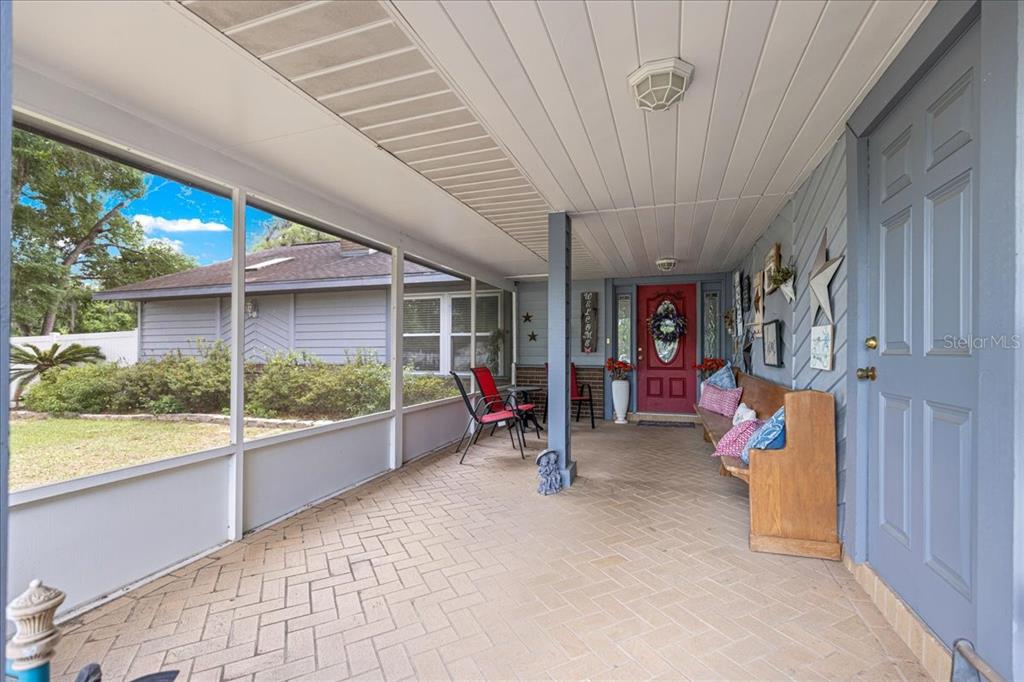 A covered porch with a brick floor, a red front door, and a wooden bench — 11470 SE 73RD COURT, BELLEVIEW, FL