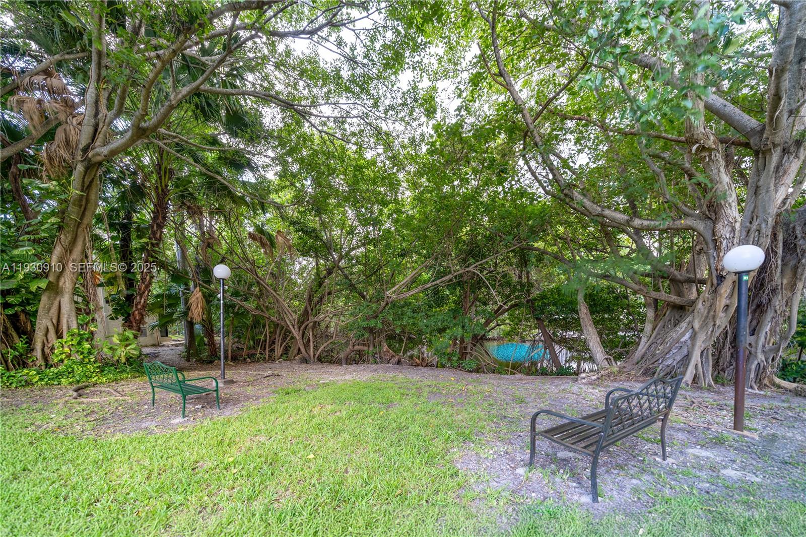 A grassy outdoor area features two metal benches, two lamp posts, and dense surrounding trees — 19212 NE 25th Ave # 283D, Miami FL 33180, MIAMI, FL