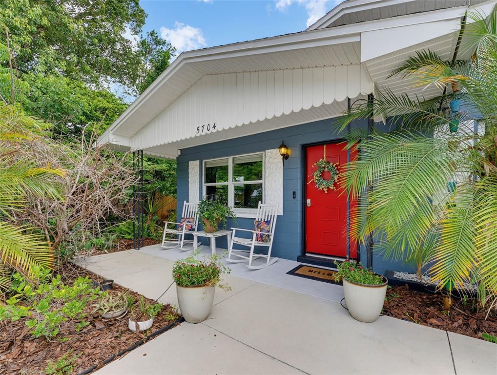 A blue house exterior features a covered porch with two rocking chairs and potted plants — 5704 13TH STREET, ZEPHYRHILLS, FL
