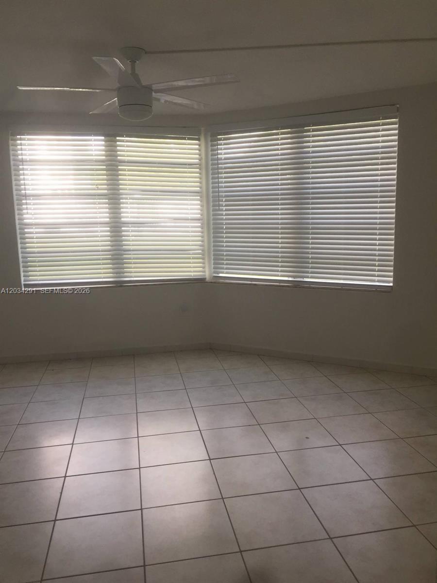 An empty room features white tile flooring, a ceiling fan, and large windows with blinds — 1351 NE 191st St # E117, Miami FL 33179, MIAMI, FL