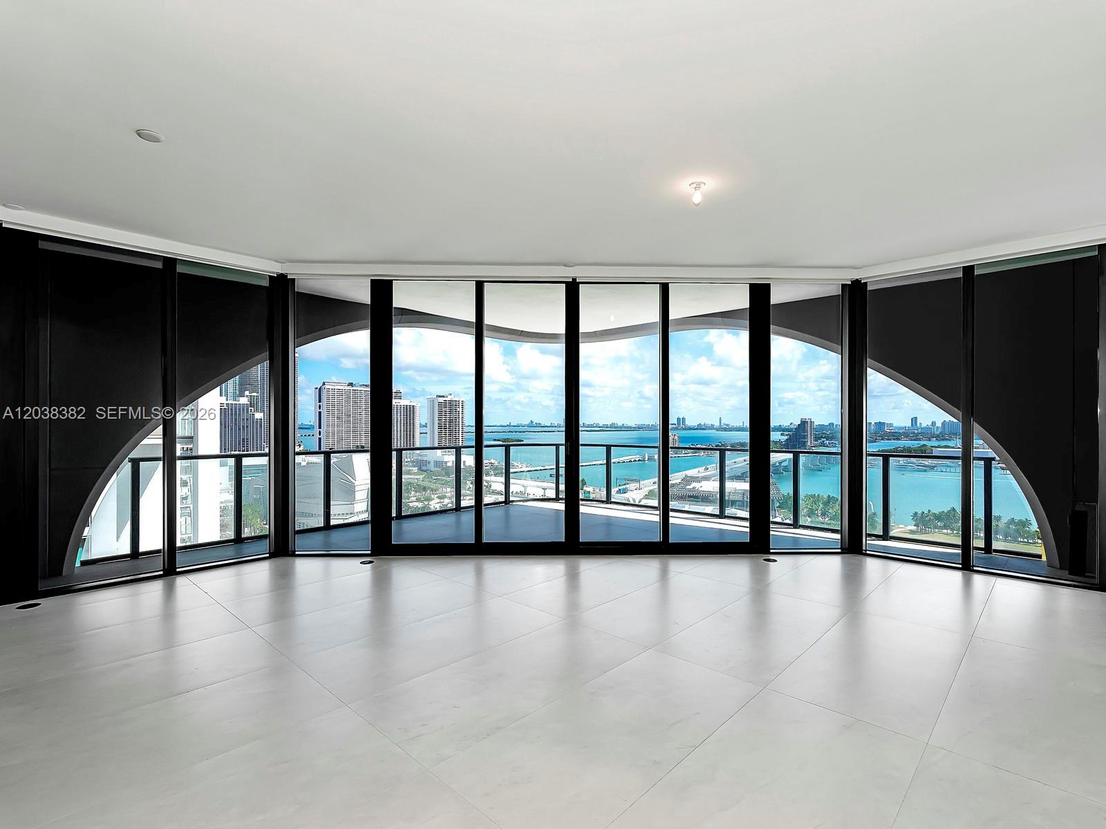 An empty room with white tile floors features floor-to-ceiling windows overlooking a bay and city — 1000 Biscayne Blvd # 2002, Miami FL 33132, MIAMI, FL