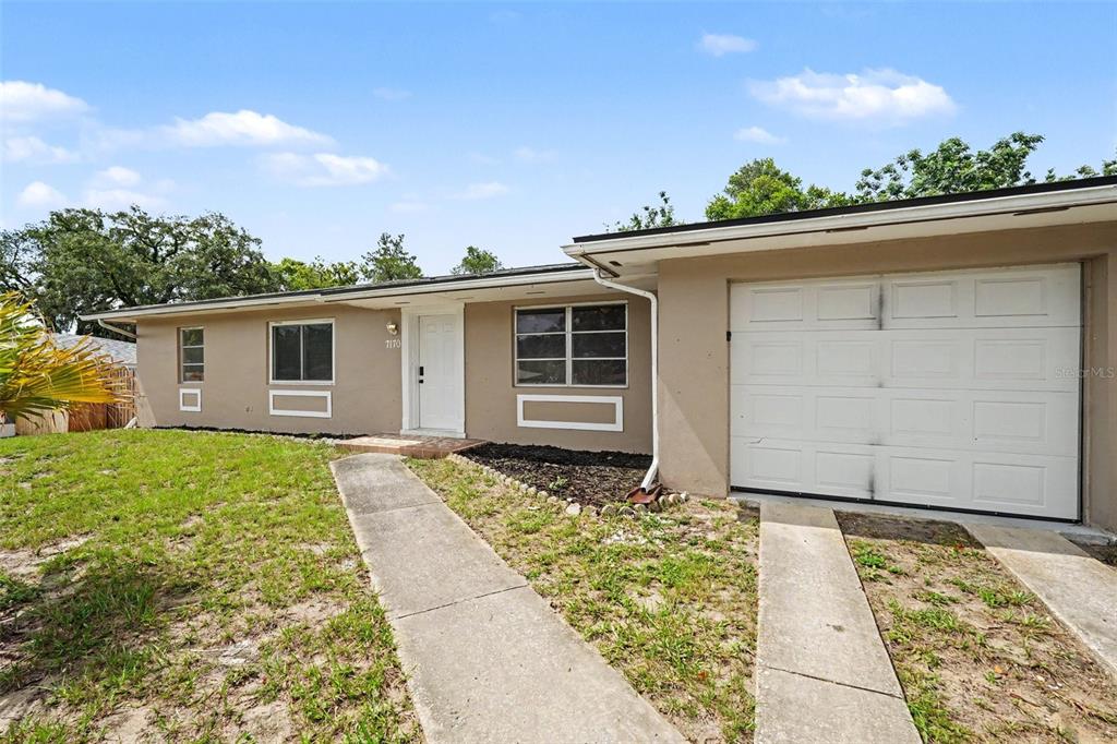 A single-story house with a beige exterior, a white garage door, and a concrete walkway — 7170 HOLIDAY DRIVE, SPRING HILL, FL