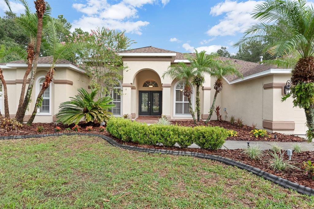 A single-story stucco house with a front entryway, palm trees, and landscaped garden beds — 5711 TANAGERSIDE ROAD, LITHIA, FL