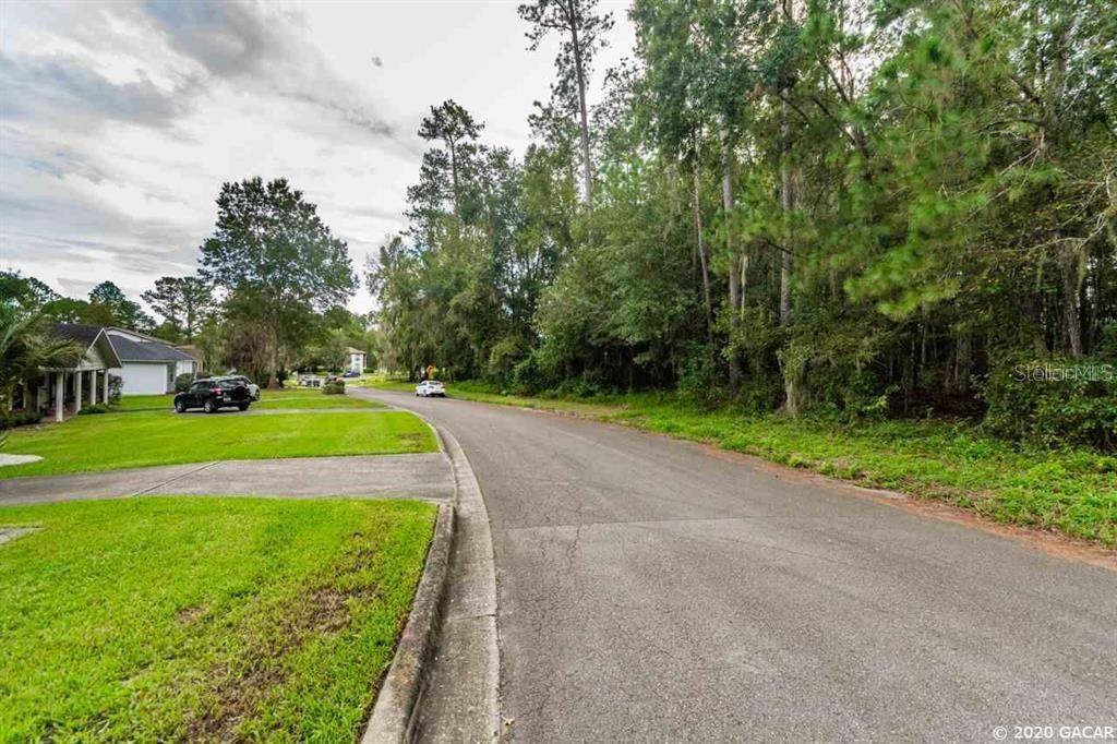 A paved residential street curves past a grassy lawn and a dense wooded area — 2804 NW 104TH COURT, GAINESVILLE, FL