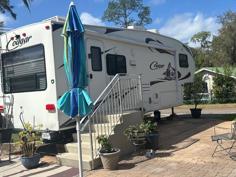 A white Cougar fifth-wheel RV is parked on a paved patio with stairs and an umbrella — 14114 252ND TERRACE ROAD NE LOT 12, FORT MC COY, FL