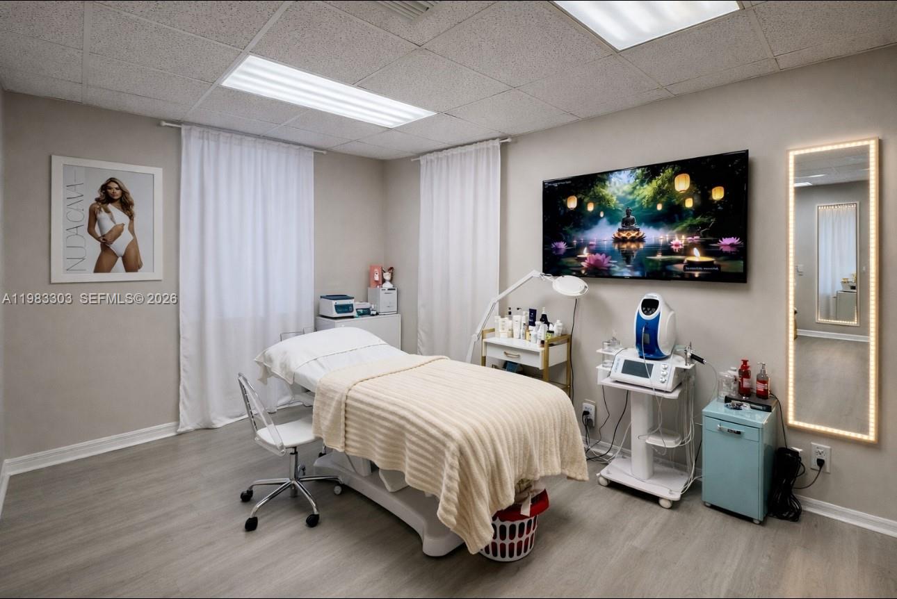 This treatment room features a massage table, medical equipment, a wall-mounted television, and a mirror — 2100 E Hallandale Beach Blvd, Hallandale Beach FL 33009, HALLANDALE BEACH, FL