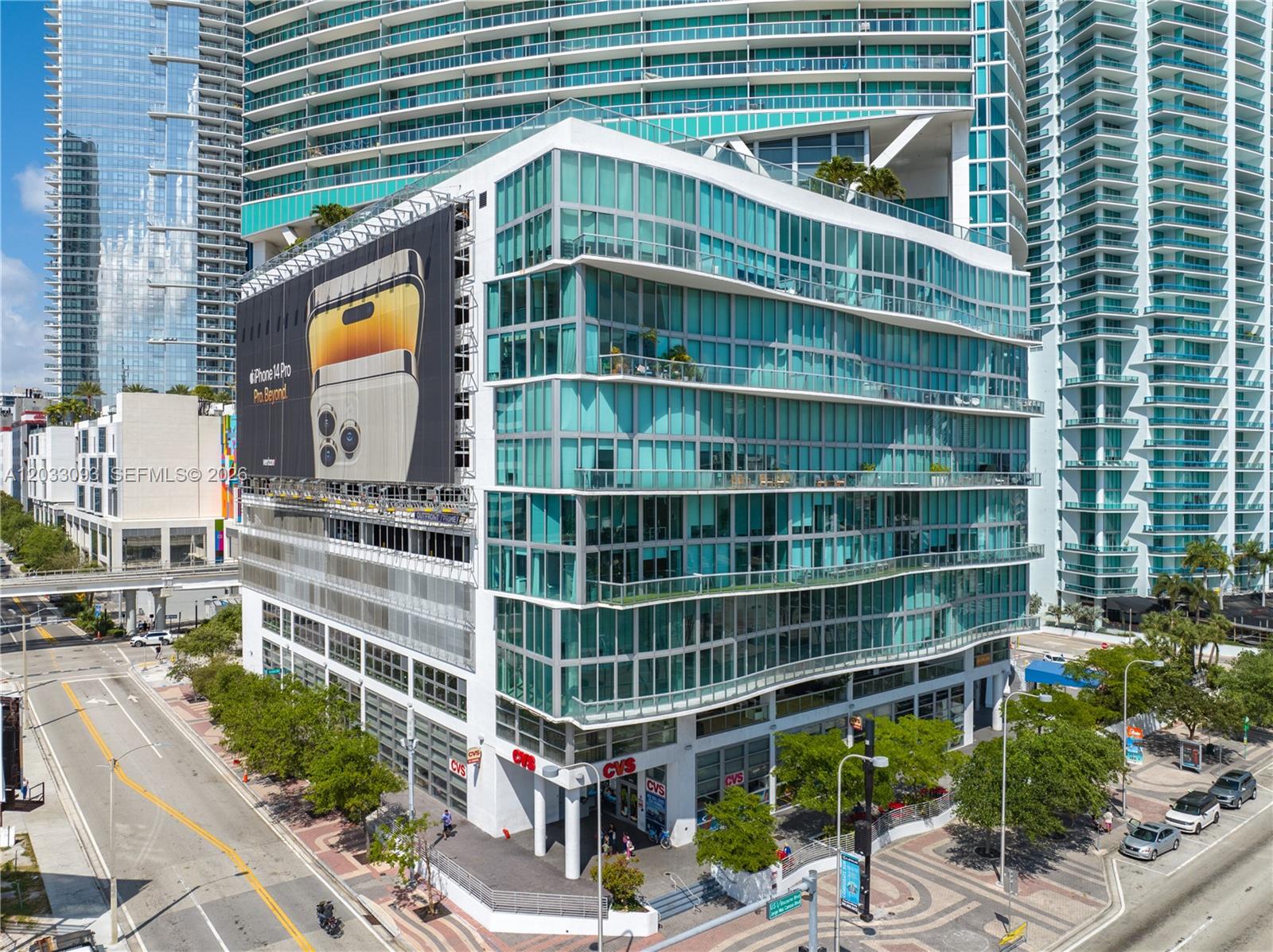 An aerial view of a modern glass-facade building with a large advertisement and ground-level retail — 888 Biscayne Blvd, Miami FL 33132, MIAMI, FL