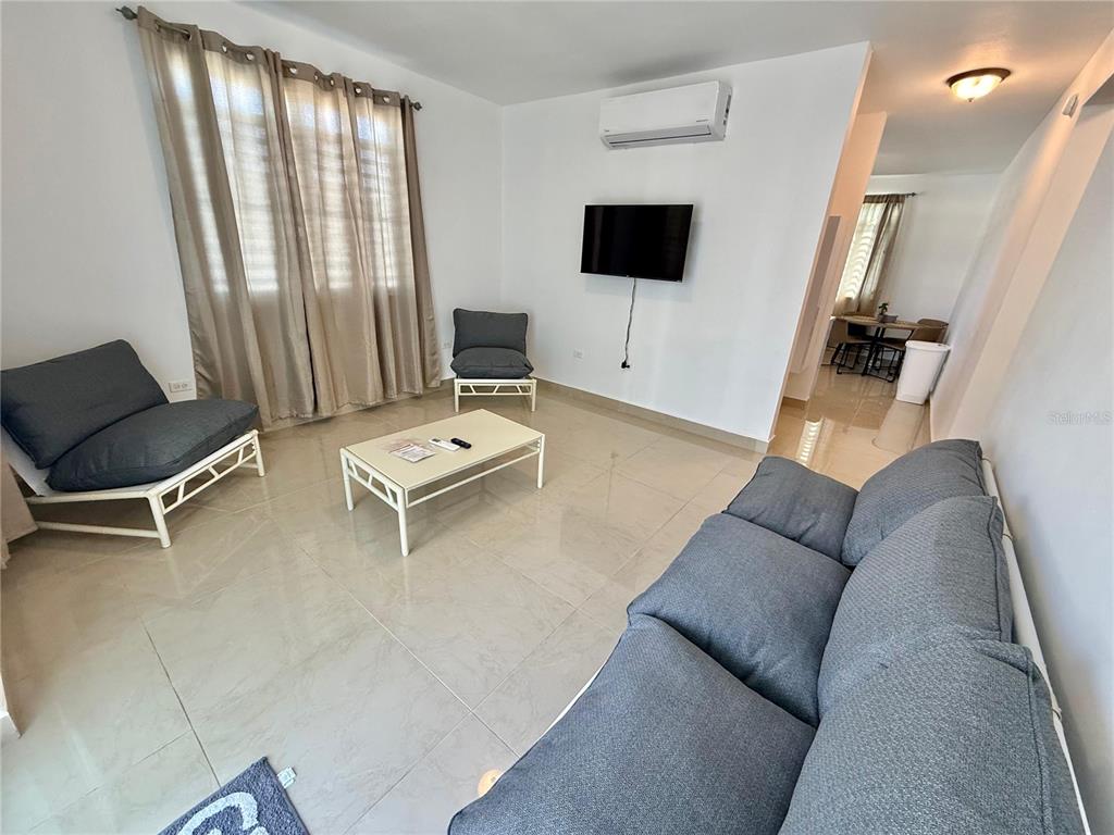A living room features light tile flooring, gray furniture, a wall-mounted television, and beige curtains — Carr 115 km 8.7 CALLE HOSTOS, RINCON, PR