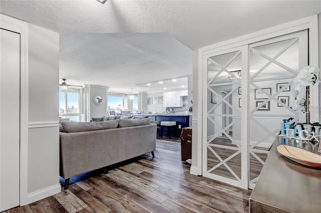 A living room with wood-look flooring leads to a kitchen featuring white cabinets and blue accents — 644 ISLAND WAY UNIT 101, CLEARWATER BEACH, FL