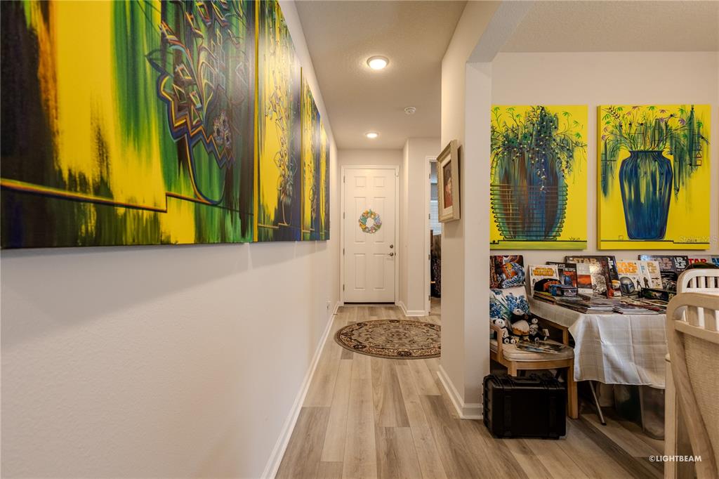 A hallway with light wood flooring leads to a white front door with artwork displayed — 10297 CHURCH HAMMOCK ROAD, LEESBURG, FL