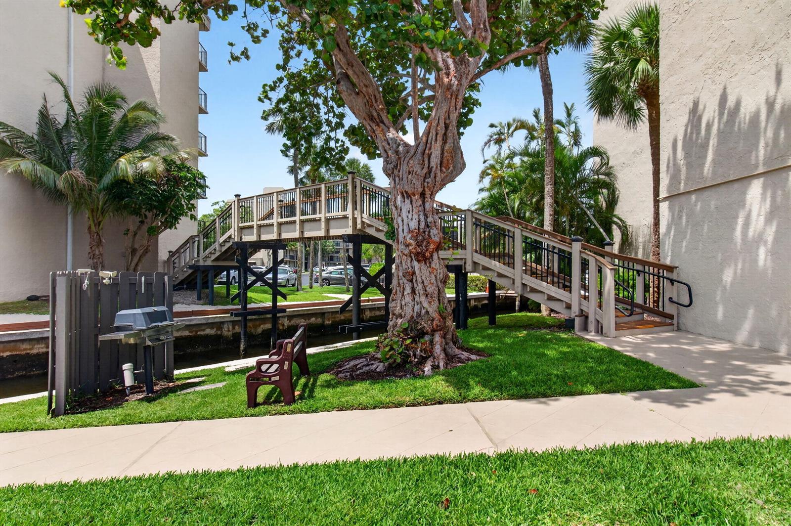 A wooden pedestrian bridge crosses a canal between two apartment buildings near a large tree — 12 Royal Palm Way # 501, Boca Raton FL 33432, BOCA RATON, FL