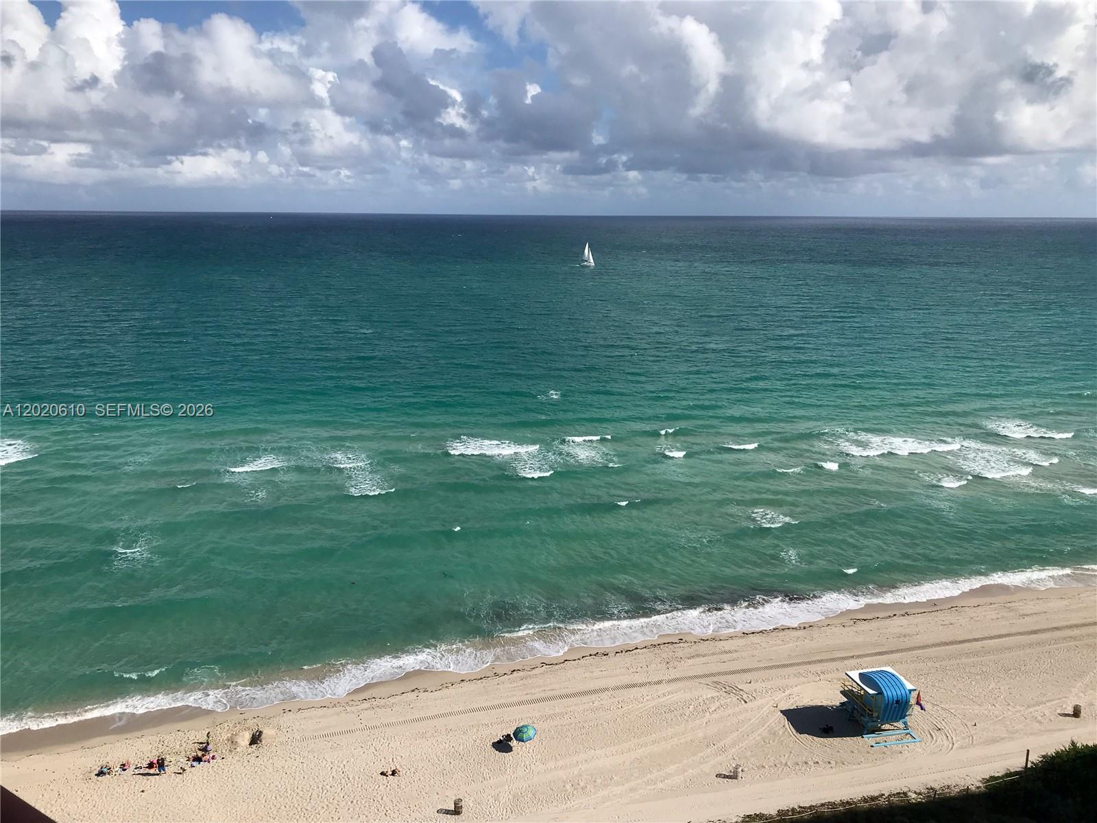 An elevated view of a sandy beach, turquoise ocean, and a single sailboat under clouds — MIAMI BEACH, FL