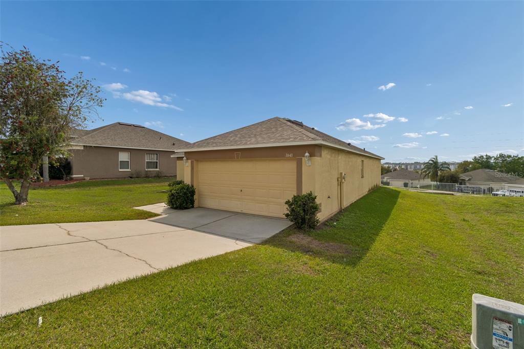 A tan single-story house with a two-car garage sits on a grassy corner lot — 7643 WEEHAWKEN DRIVE, ZEPHYRHILLS, FL