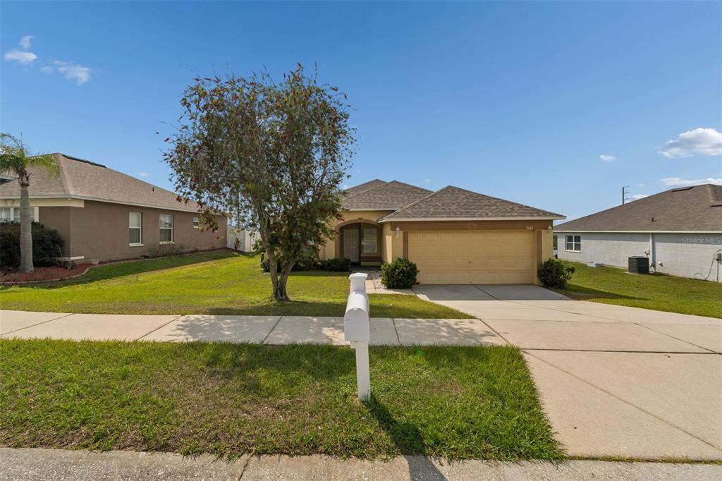 A single-story suburban house with a tan stucco exterior, a two-car garage, and a front lawn — 7643 WEEHAWKEN DRIVE, ZEPHYRHILLS, FL