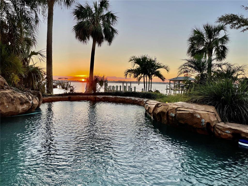 A swimming pool with rock borders overlooks a waterfront sunset with palm trees and docks — 2904 S PENINSULA DR, DAYTONA BEACH SHORES, FL