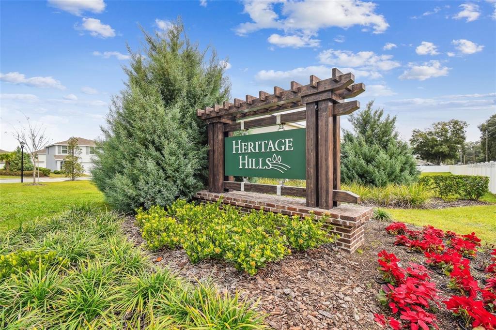 A wooden sign for Heritage Hills stands in a landscaped garden with red flowers — 13688 WINEBERRY DRIVE, DADE CITY, FL