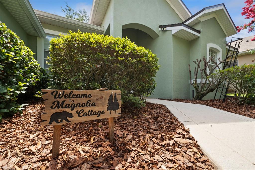 A wooden sign reading Welcome to Magnolia Cottage sits in front of a green house — 2243 WOLF RIDGE LANE, MOUNT DORA, FL