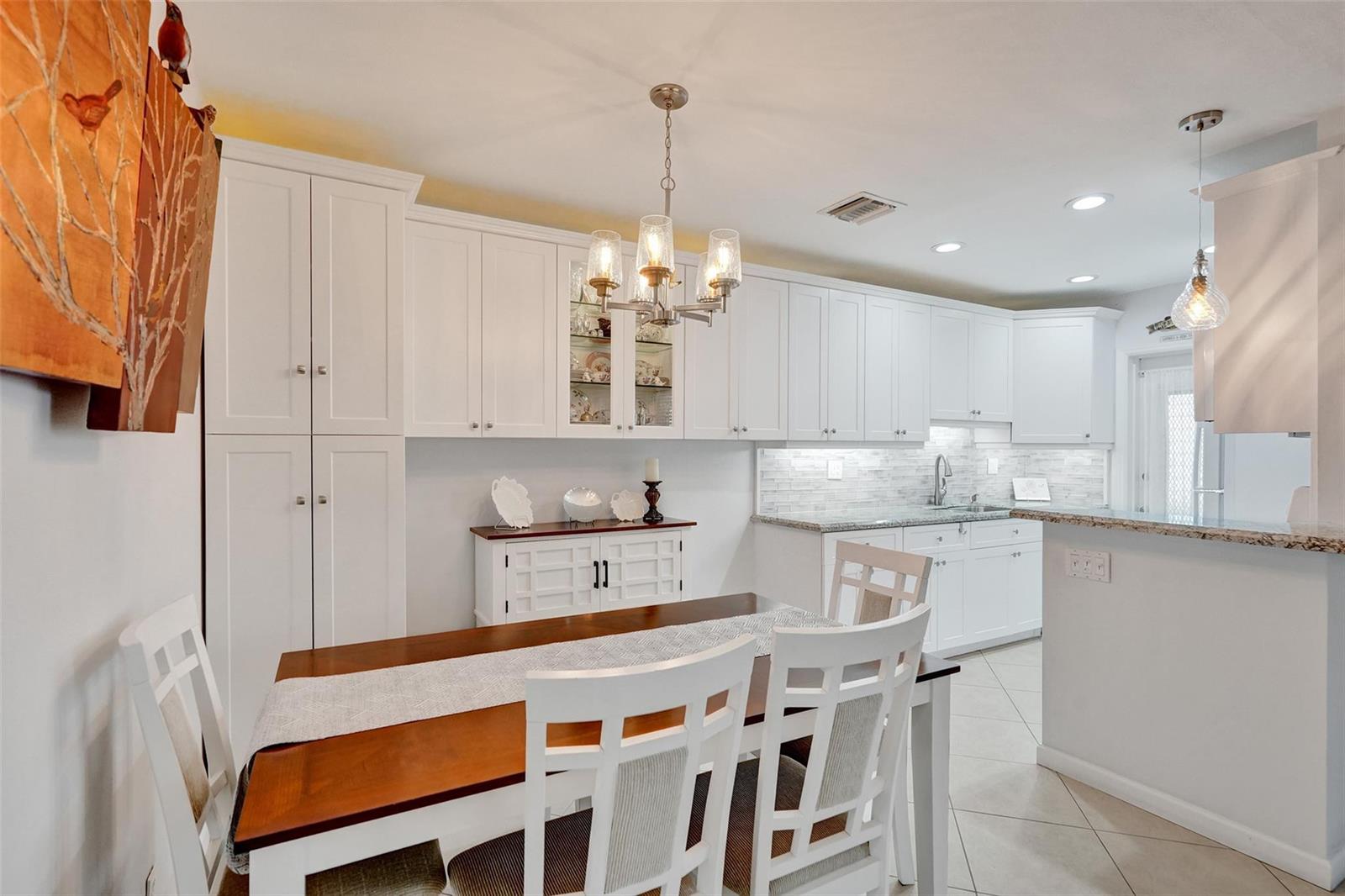 A dining area with a wooden table and white chairs sits adjacent to a white kitchen — 935 SE 9th Avenue # 16, Pompano Beach FL 33060, POMPANO BEACH, FL