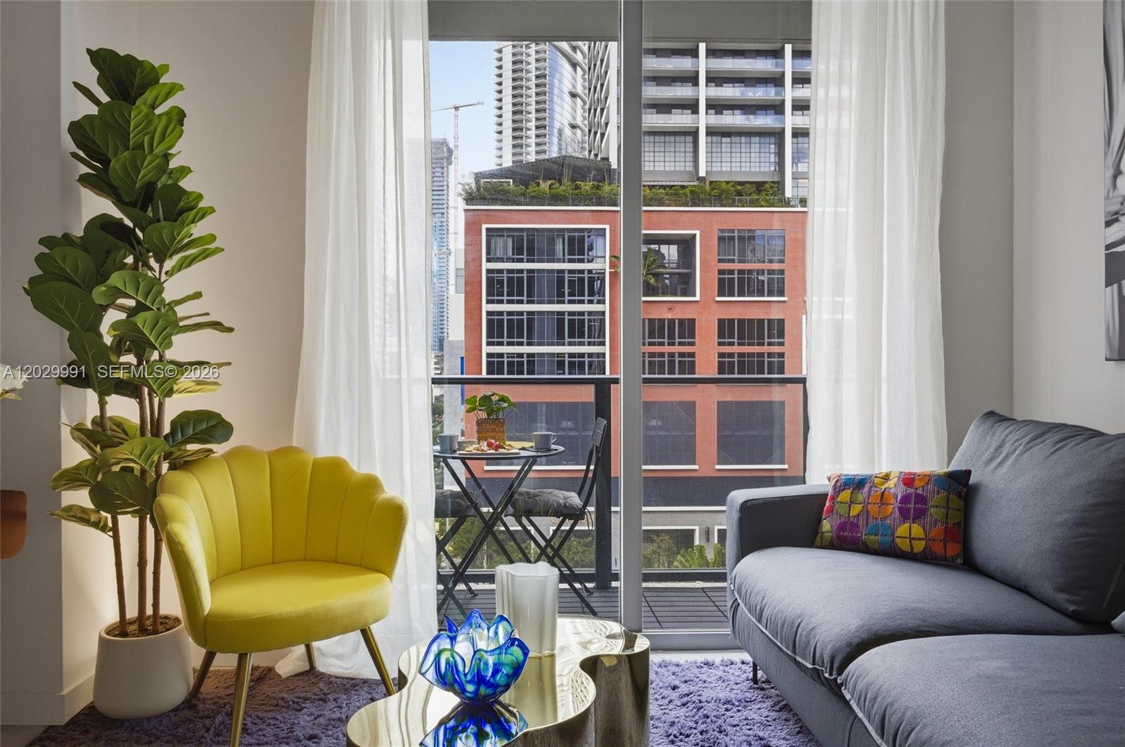 A living room features a yellow chair, gray sofa, and balcony with a city view — 121 NE 5th St # 711, Miami FL 33132, MIAMI, FL