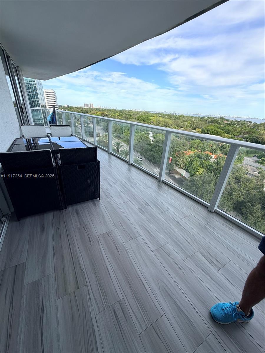 A balcony features wood-look tile flooring, a dining set, and a glass railing overlooking greenery — 4250 Biscayne Blvd # 902, Miami FL 33137, MIAMI, FL