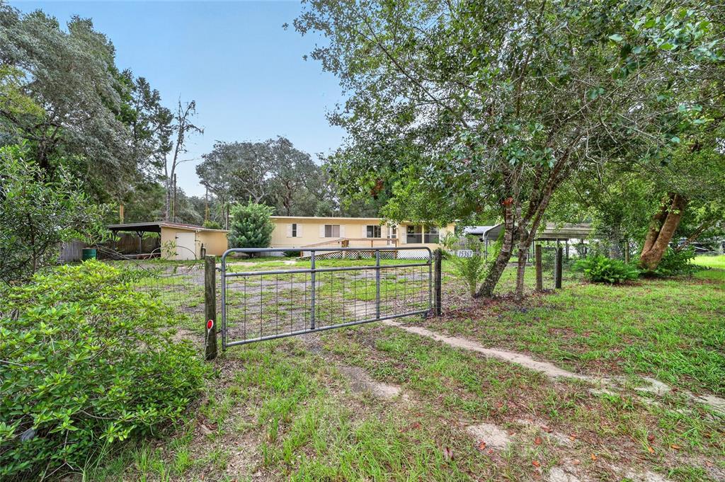 A metal gate opens to a yard with a single-story house and a detached carport — 15367 NE 160TH AVENUE ROAD, FORT MC COY, FL