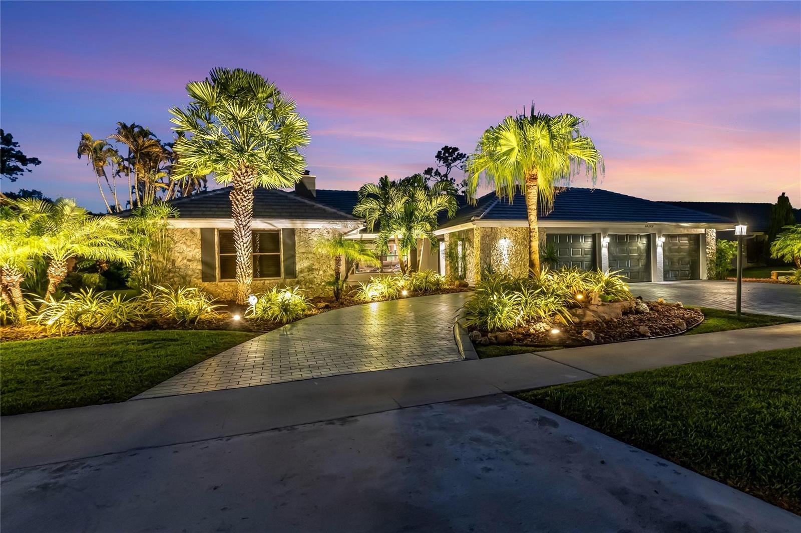 A stone-faced house with a paved driveway and palm trees at dusk — 10182 Boca Woods Lane, Boca Raton FL 33428, BOCA RATON, FL