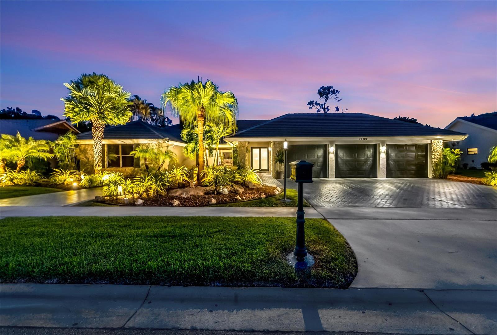 A single-story house with a three-car garage and illuminated landscaping at dusk — 10182 Boca Woods Lane, Boca Raton FL 33428, BOCA RATON, FL