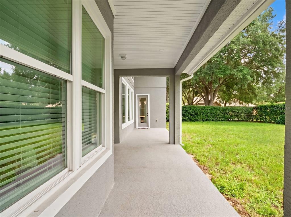 A covered concrete patio overlooks a grassy backyard with a hedge and trees — 25622 HAWKS RUN LANE, SORRENTO, FL